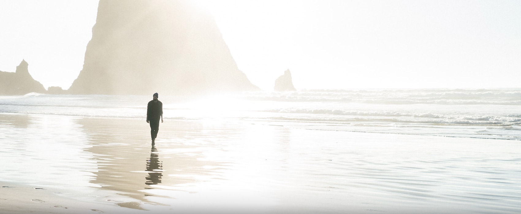 Silhuette eines Mannes, der am Strand geht, im Hintergrund ist ein Felsen zu sehen, es ist dunstig.