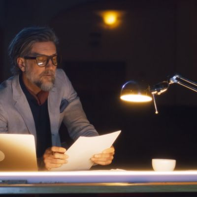 Lesender Mann mit Brille sitzt am Schreibtisch bei schwacher Beleuchtung.